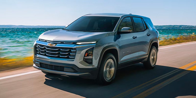 A silver blue 2026 Chevrolet Equinox being driven on a highway beside a deep aquamarine body of water.below the blue sky.
